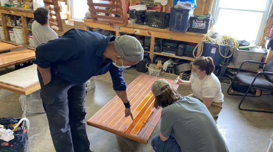 People in a workshop refinishing a wooden panel together.